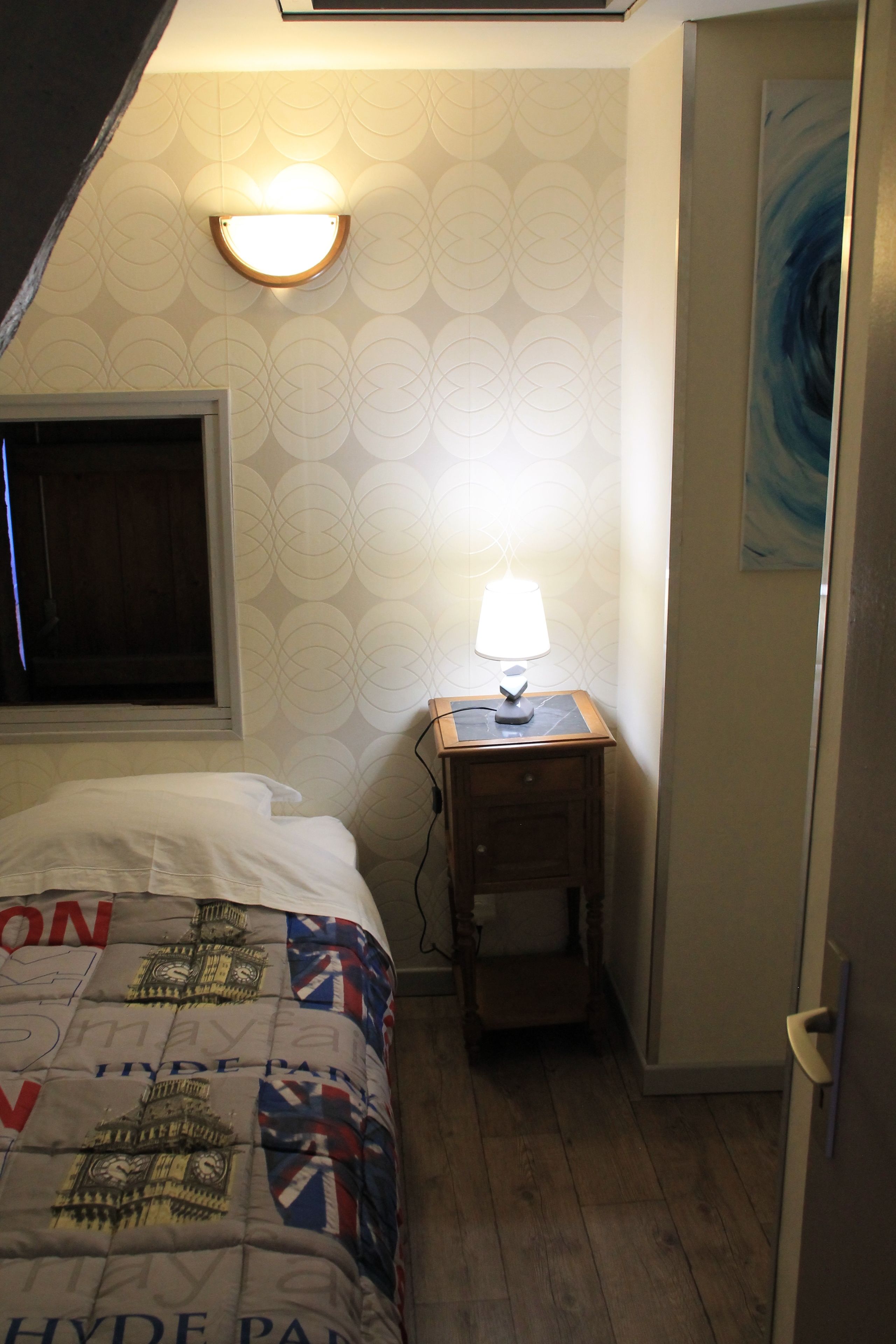 Un dormitorio con cama, mesita de noche con lámpara, ventana y un cuadro en la pared.