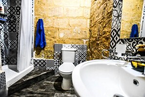 A bathroom with a white sink, a toilet, and a shower area. The walls are tiled with black and white patterns, and there is a blue towel hanging on the wall. - Historic Townhouse in Birgu Center (Il-Birgu)
