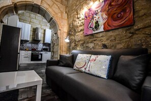 A modern living room with a sofa and a coffee table, a kitchen with white cabinets and a stone archway, and a colorful painting on the wall. - Historic Townhouse in Birgu Center (Il-Birgu)