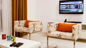 Un salon moderne avec deux canapés blancs ornés de coussins orange, une table basse blanche et un téléviseur à écran plat fixé au mur, diffusant une chaîne de sport.