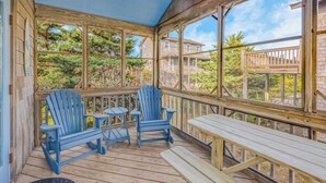 Terraza de madera con sillas Adirondack azules y una mesa de picnic, con vistas a una zona residencial con árboles y cielo despejado.