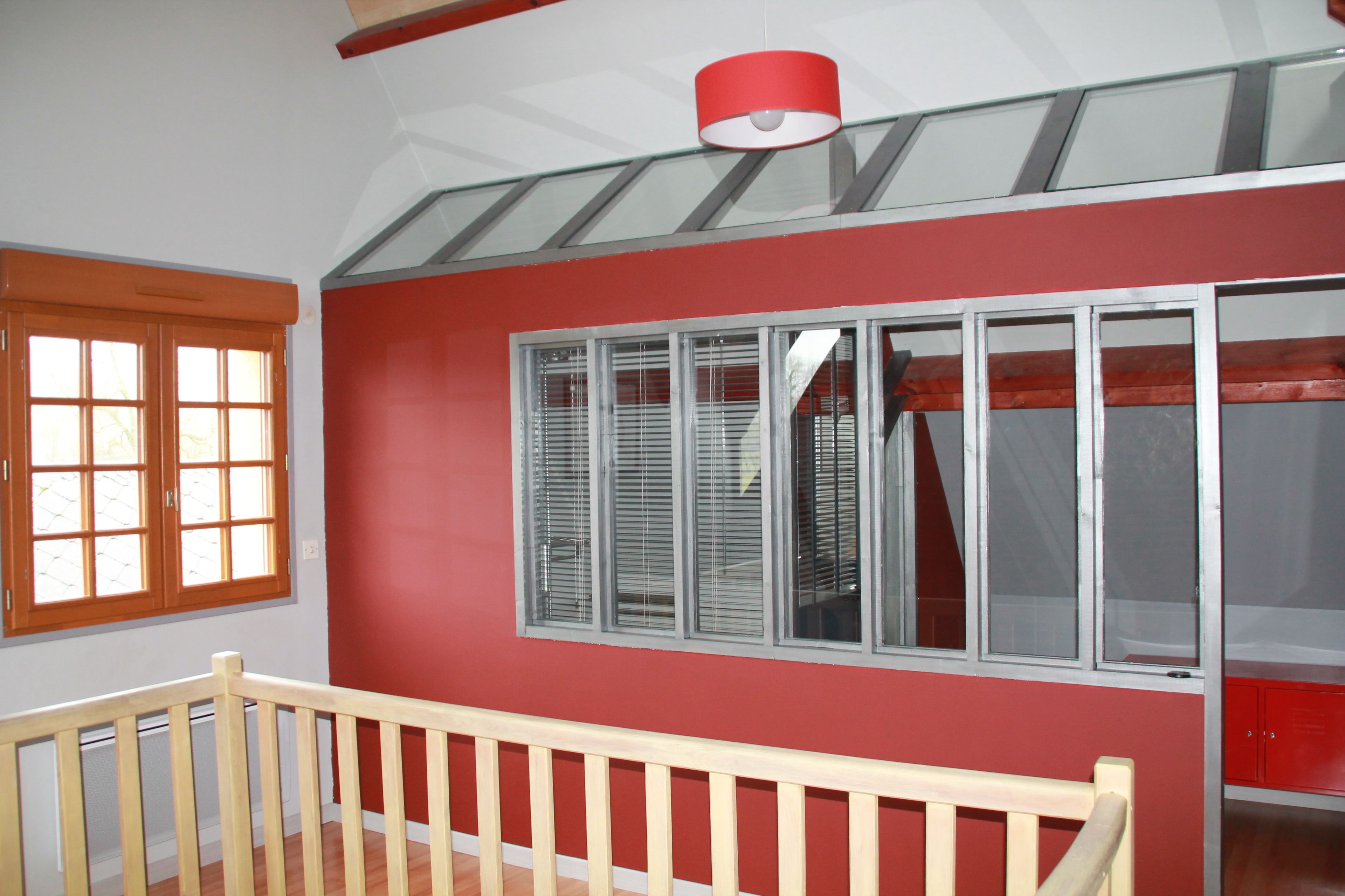 Una habitación con pared roja, un ventanal con persianas, barandal de madera y un armario rojo.