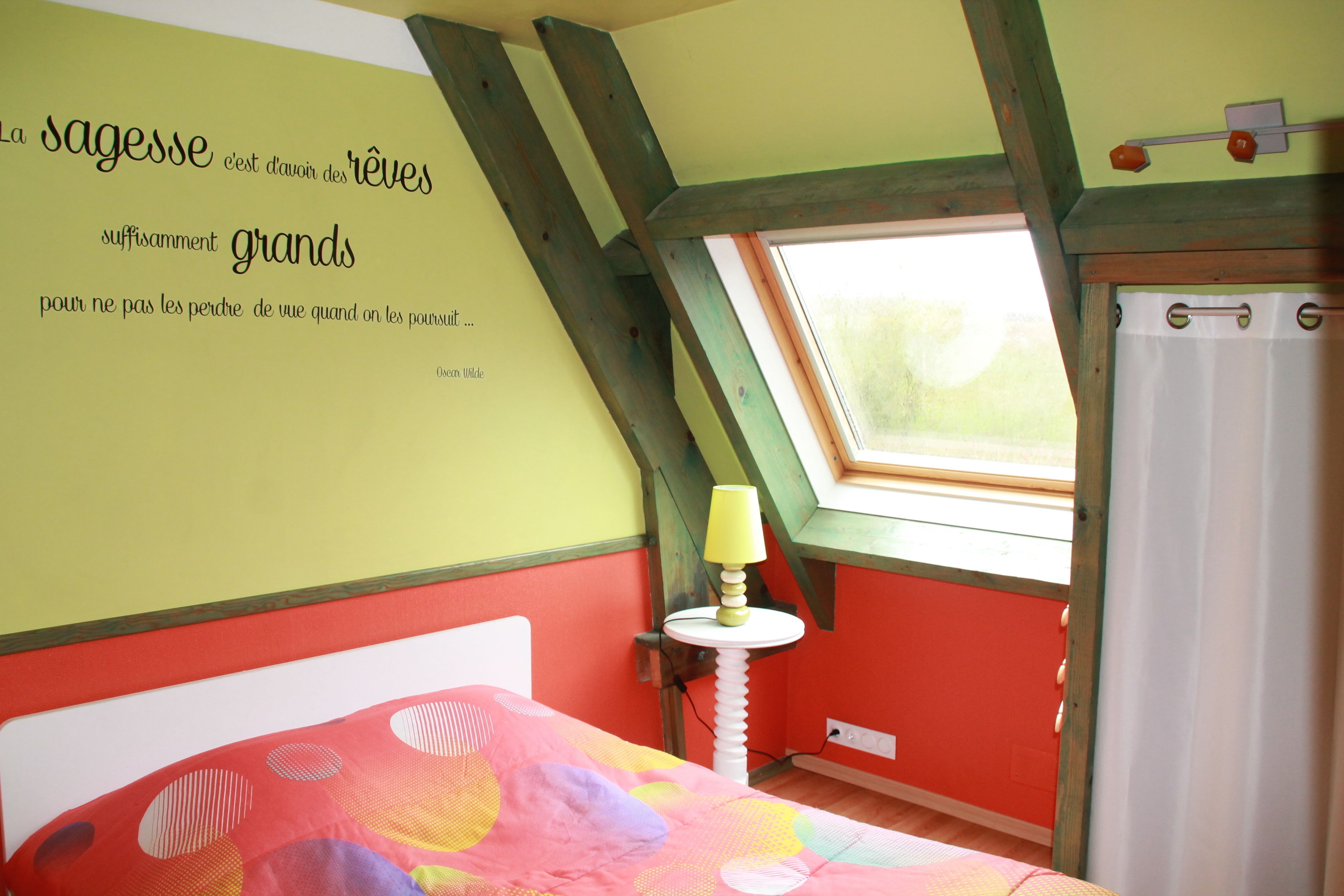 A room with a sloped ceiling, a bed with a colorful bedspread, a small table with a lamp, and a window with a curtain.