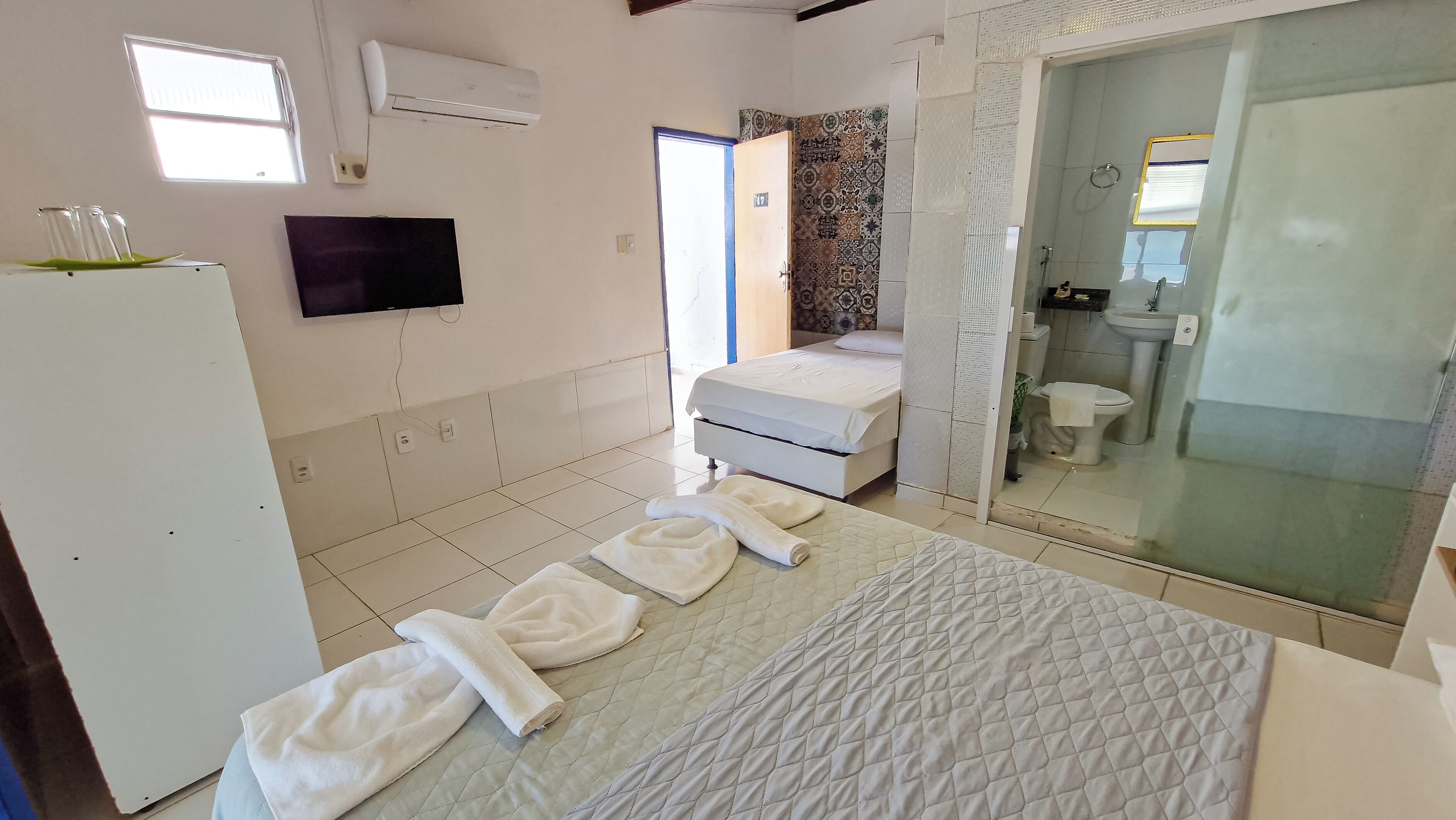 A hotel room with a bed, a TV, an air conditioner, a bathroom with a toilet and sink, and a shower area.