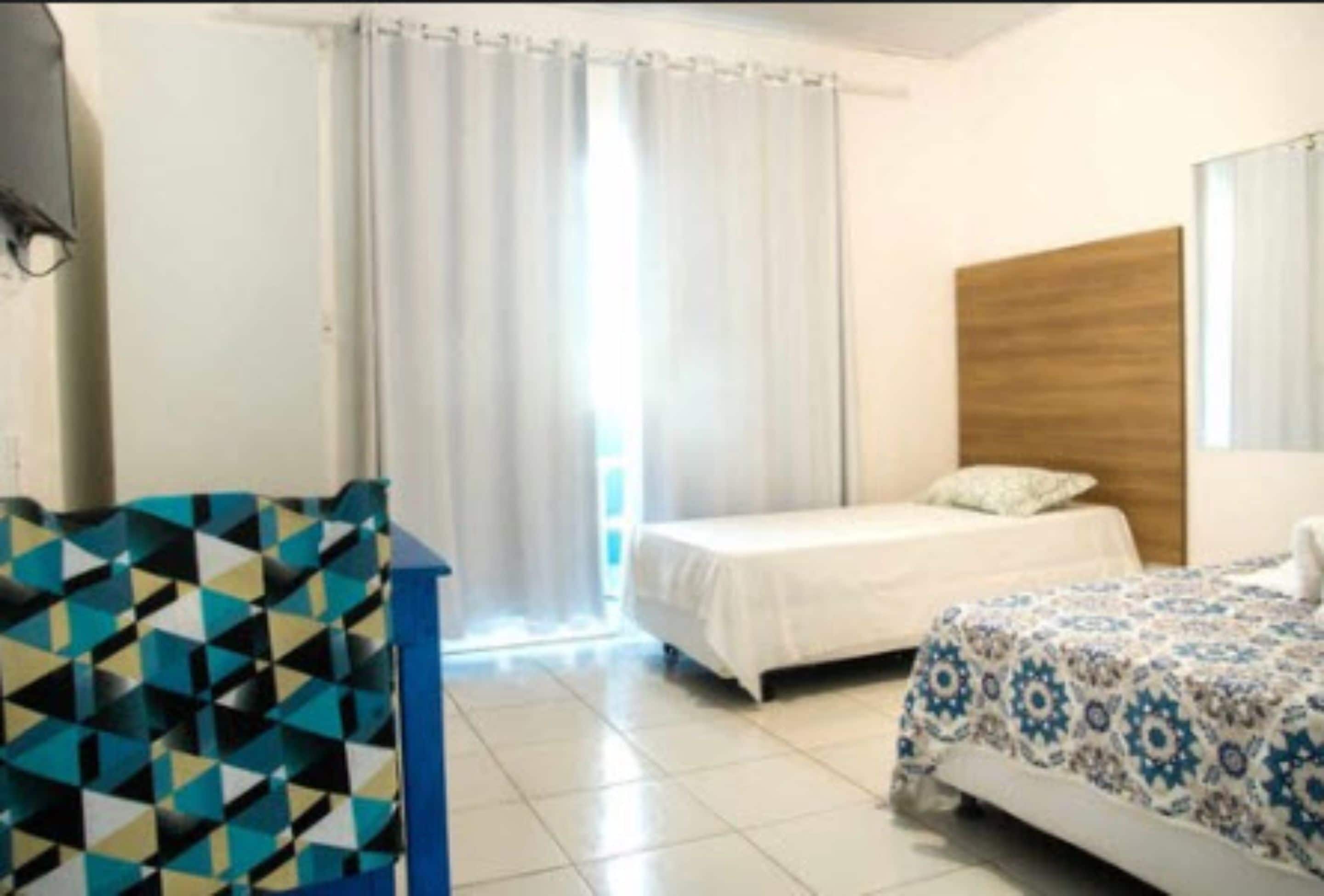 A hotel room with two beds, a wooden headboard, a large window with curtains, and a geometric-patterned chair.