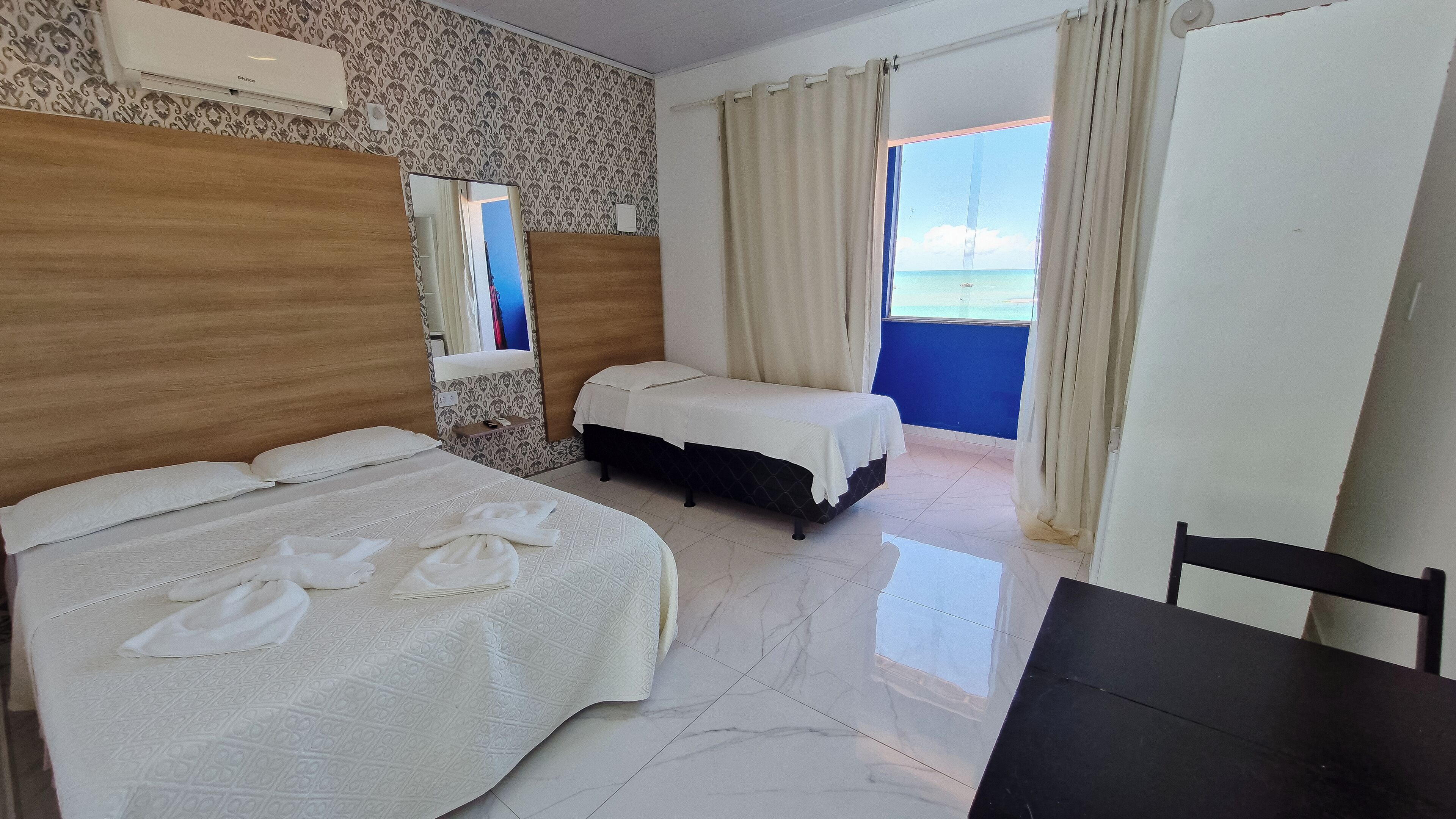 Une chambre d’hôtel avec un lit, une petite table, une chaise, un miroir et une vue sur la mer par la fenêtre.