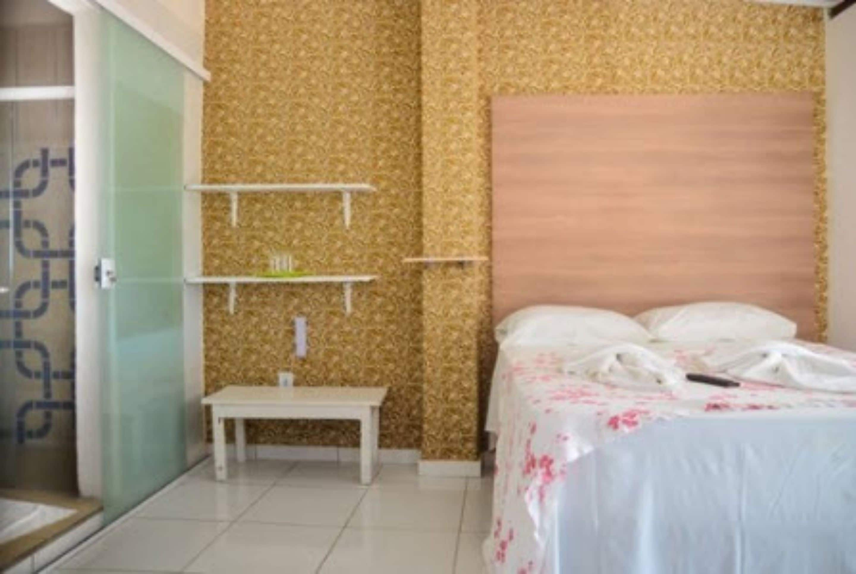 A hotel room with a bed, a bedside table, a small white table, and a shelf unit.