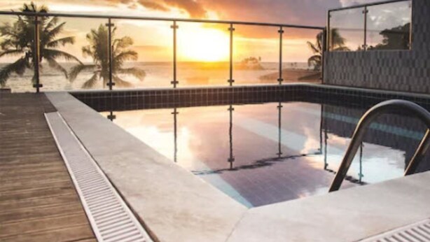 Un espace piscine offrant une vue sur le coucher de soleil, une terrasse en bois et une balustrade en verre.