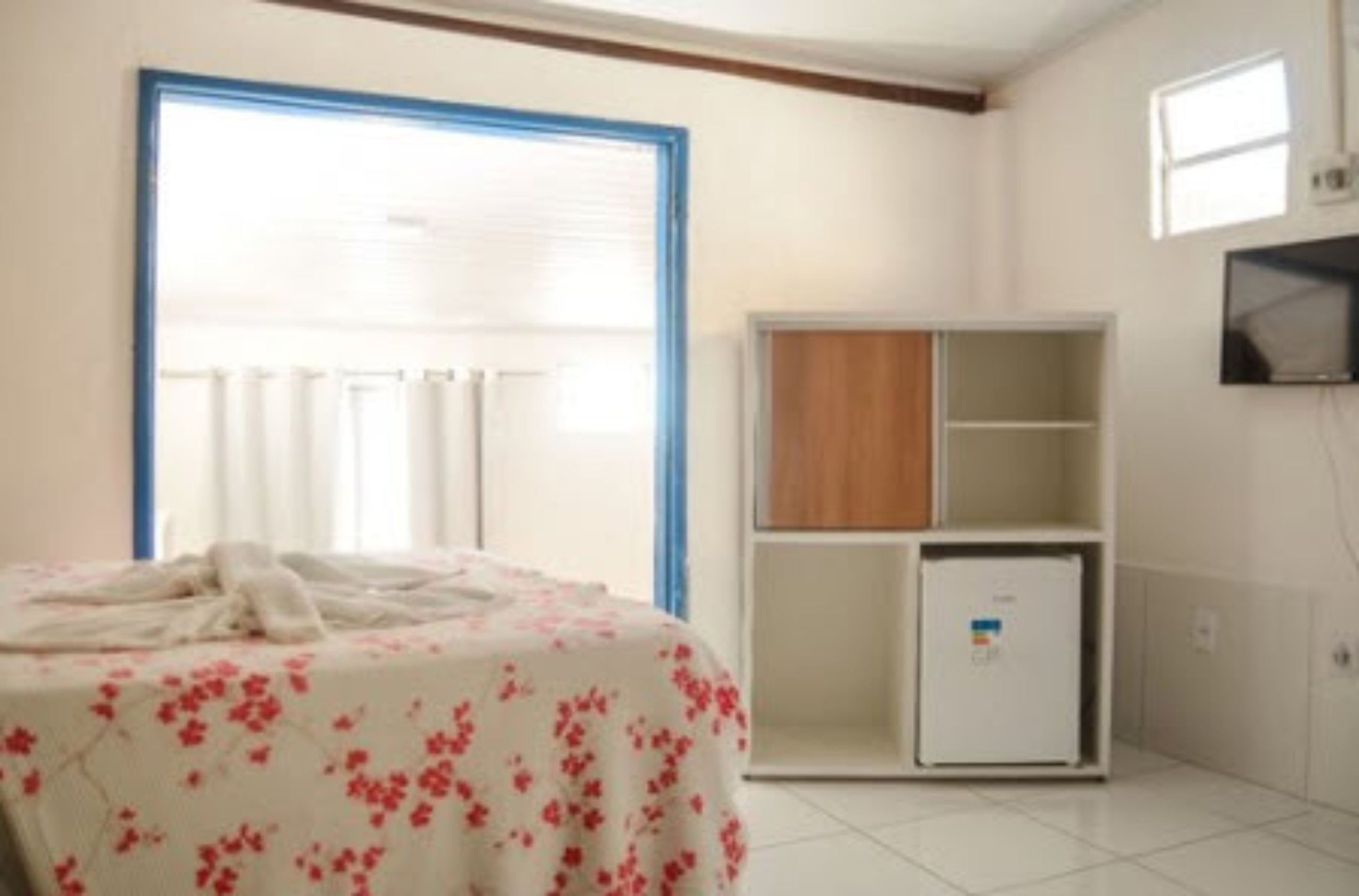 A room with a bed, a wardrobe, a small refrigerator, and a window with blinds.