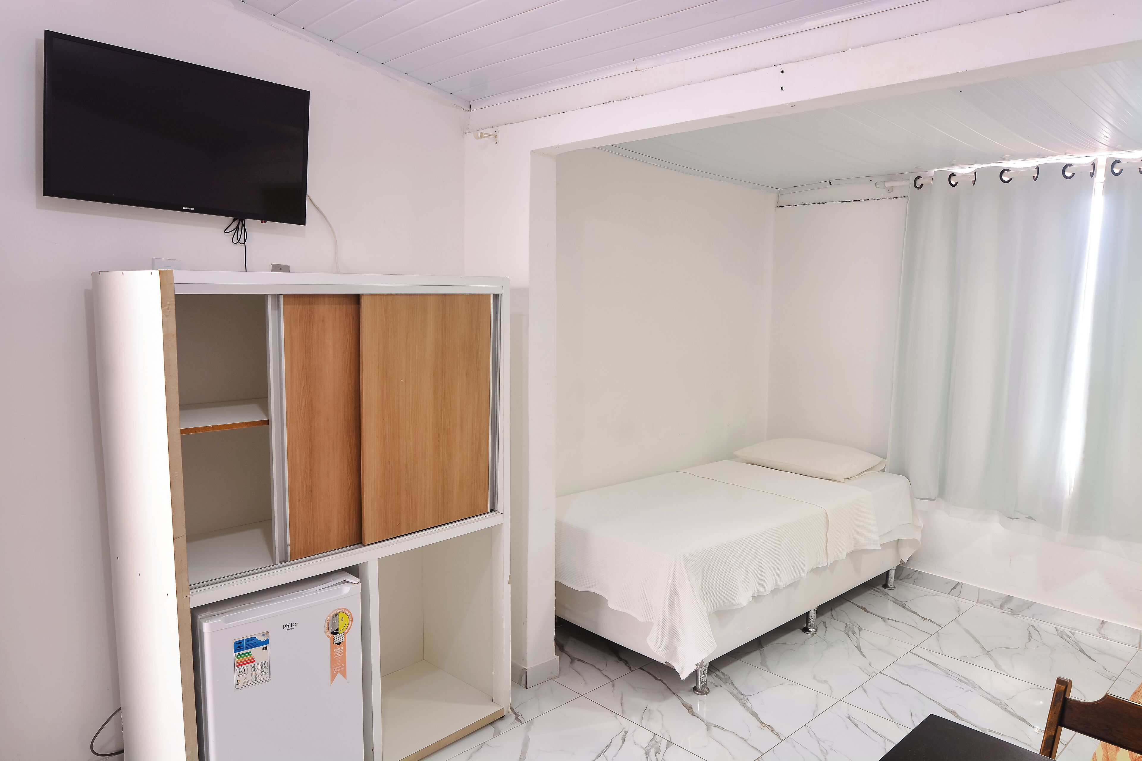 A bedroom with a bed, a TV mounted on the wall, a wardrobe, and a small refrigerator.