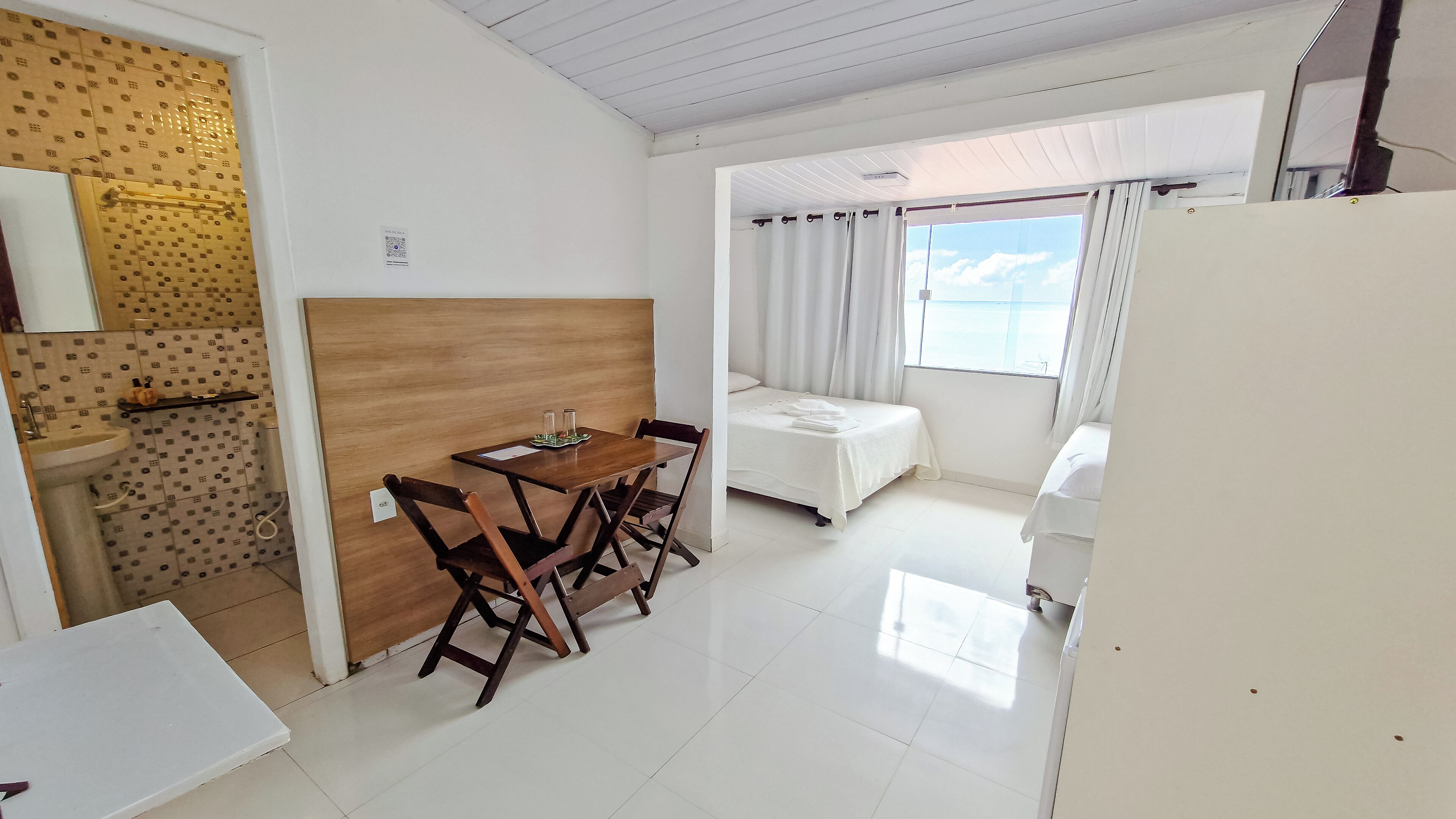 A hotel room with a wooden dining table, two chairs, a bed with white bedding, a small bathroom with a sink, and a window with curtains.
