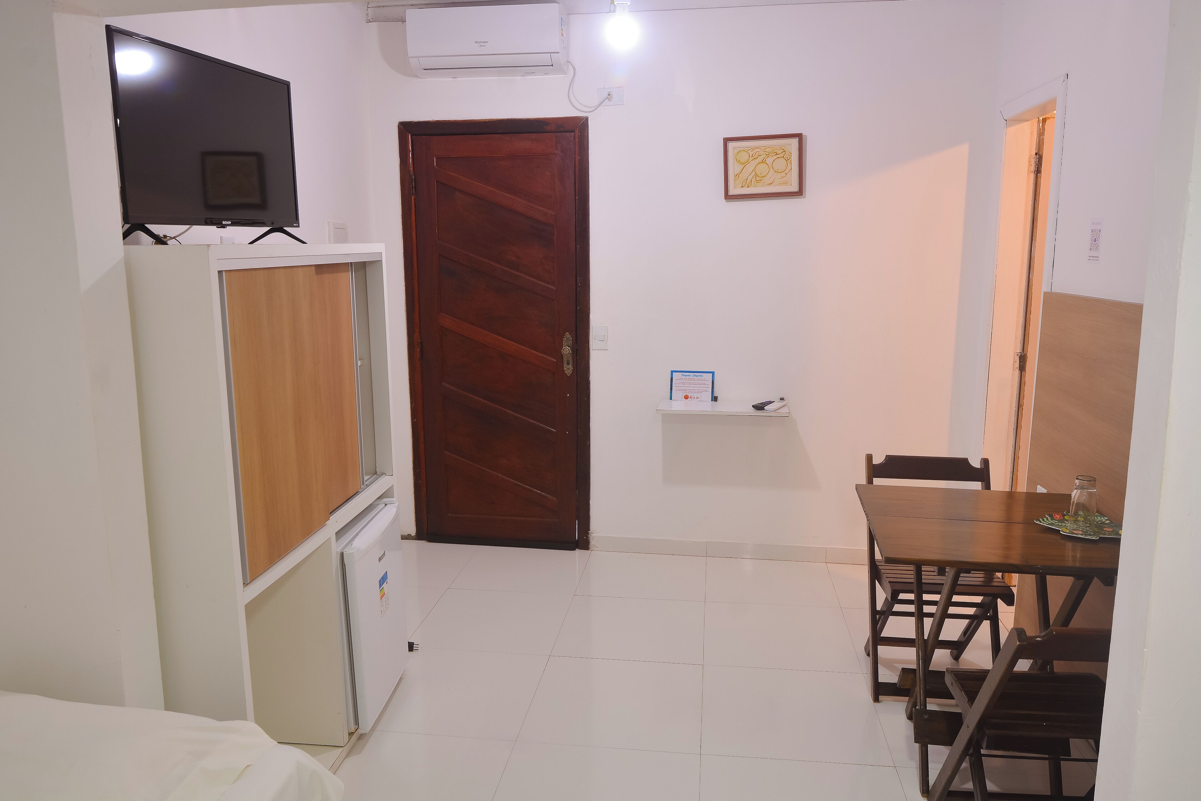 A small, clean room with a wooden door, a white cabinet, a small table with chairs, and a television mounted on the wall.