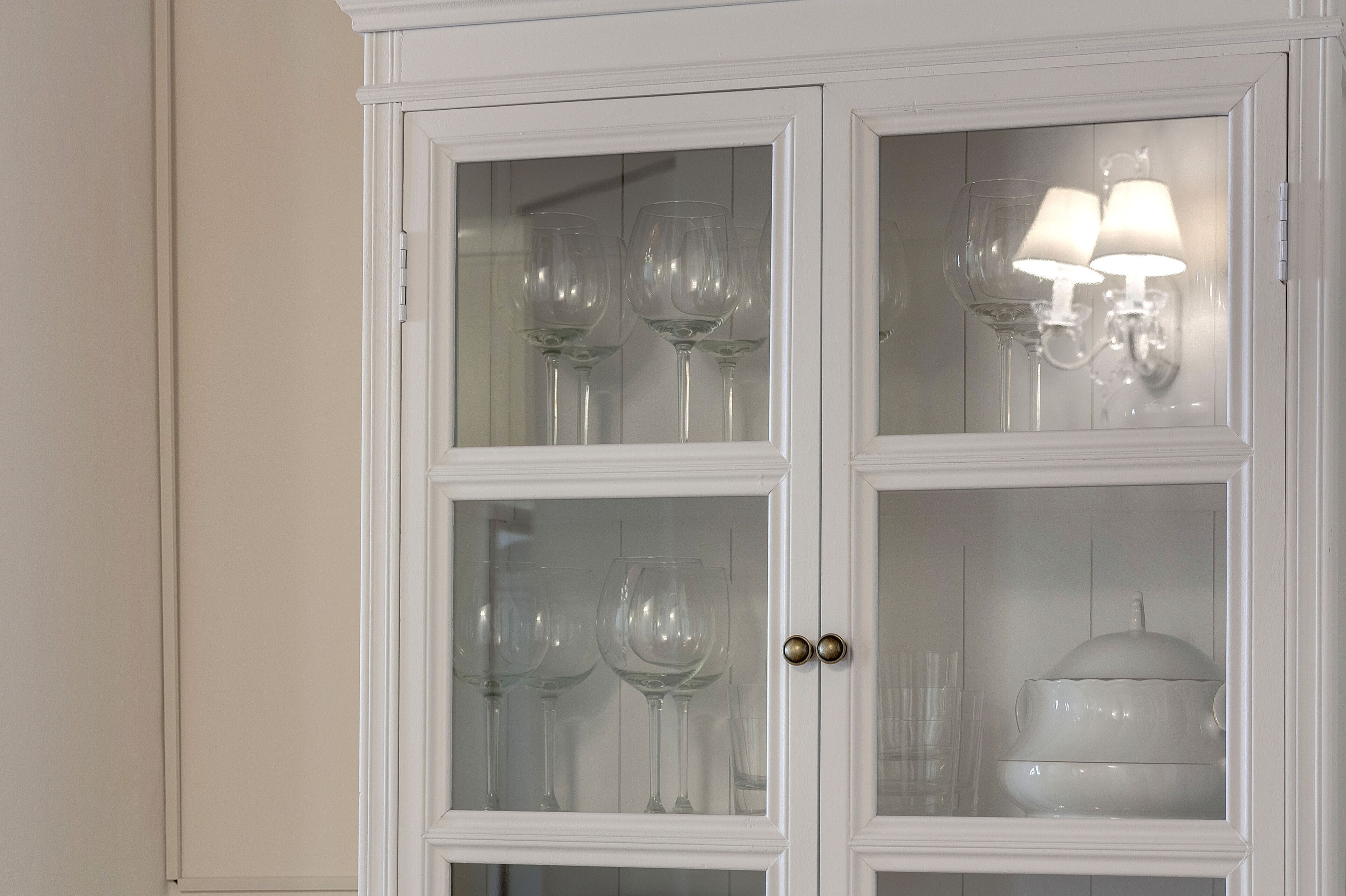 Ein weißer Schrank mit Glastüren, der Weingläser und eine Lampe beherbergt.