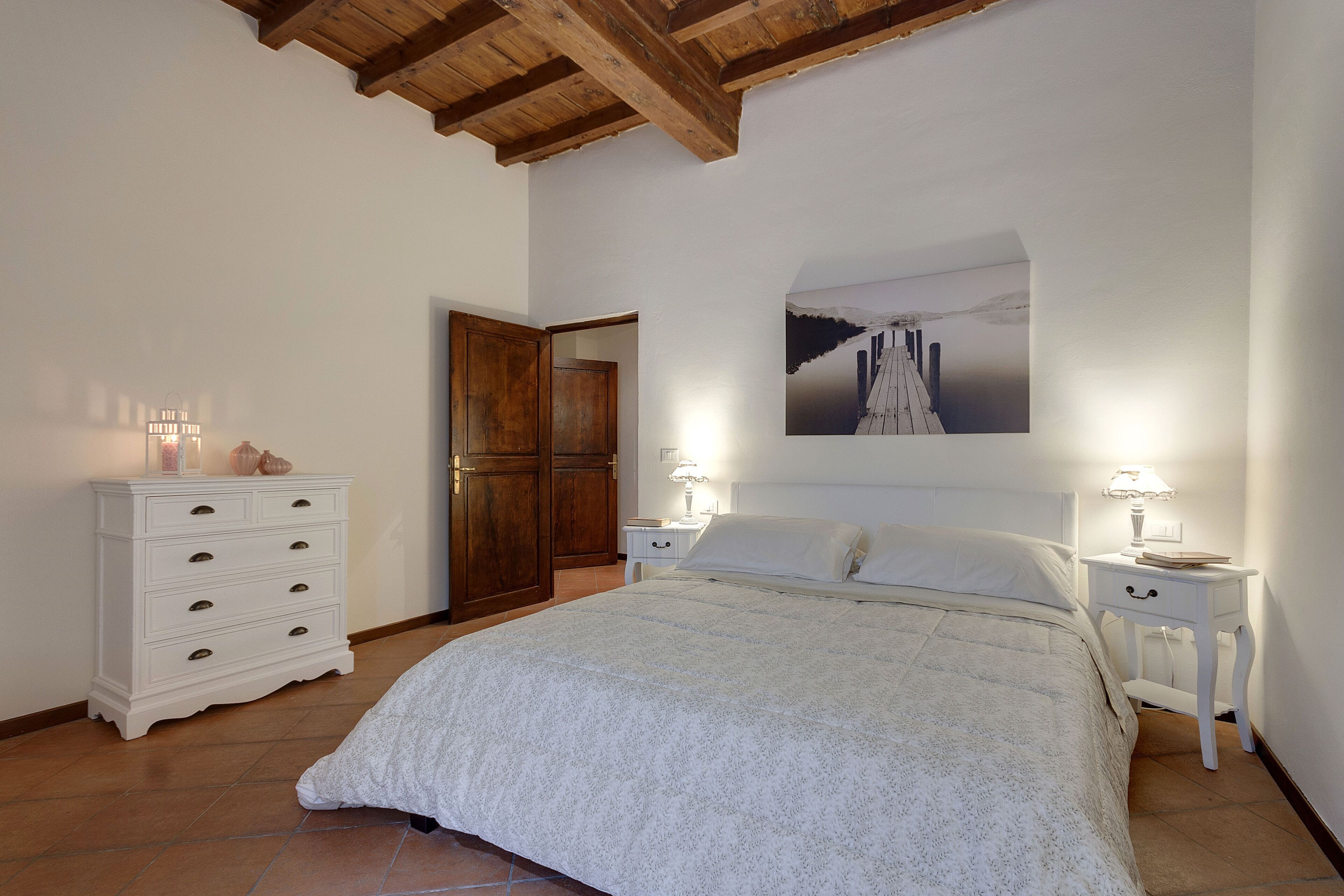 Une chambre à coucher avec un grand lit, des poutres apparentes en bois au plafond, des murs blancs, une armoire en bois et une table de chevet avec une lampe.