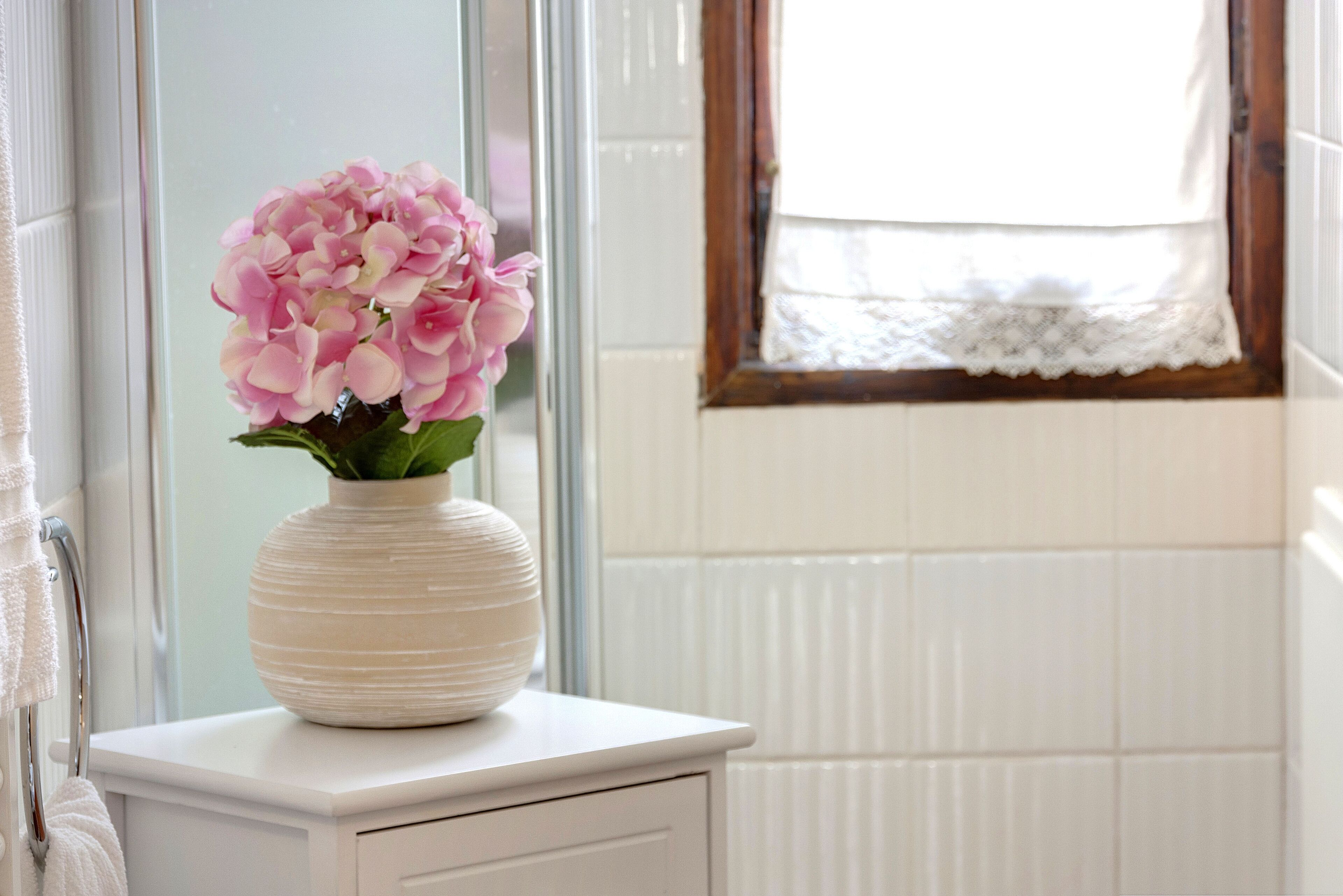 Un jarrón blanco con flores rosadas sobre un aparador blanco cerca de una ventana con cortina de encaje.