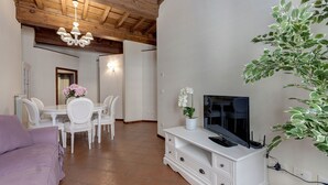 Una sala de estar con un televisor sobre un mueble blanco, un comedor con sillas y una mesa blancos, y un sofá morado.