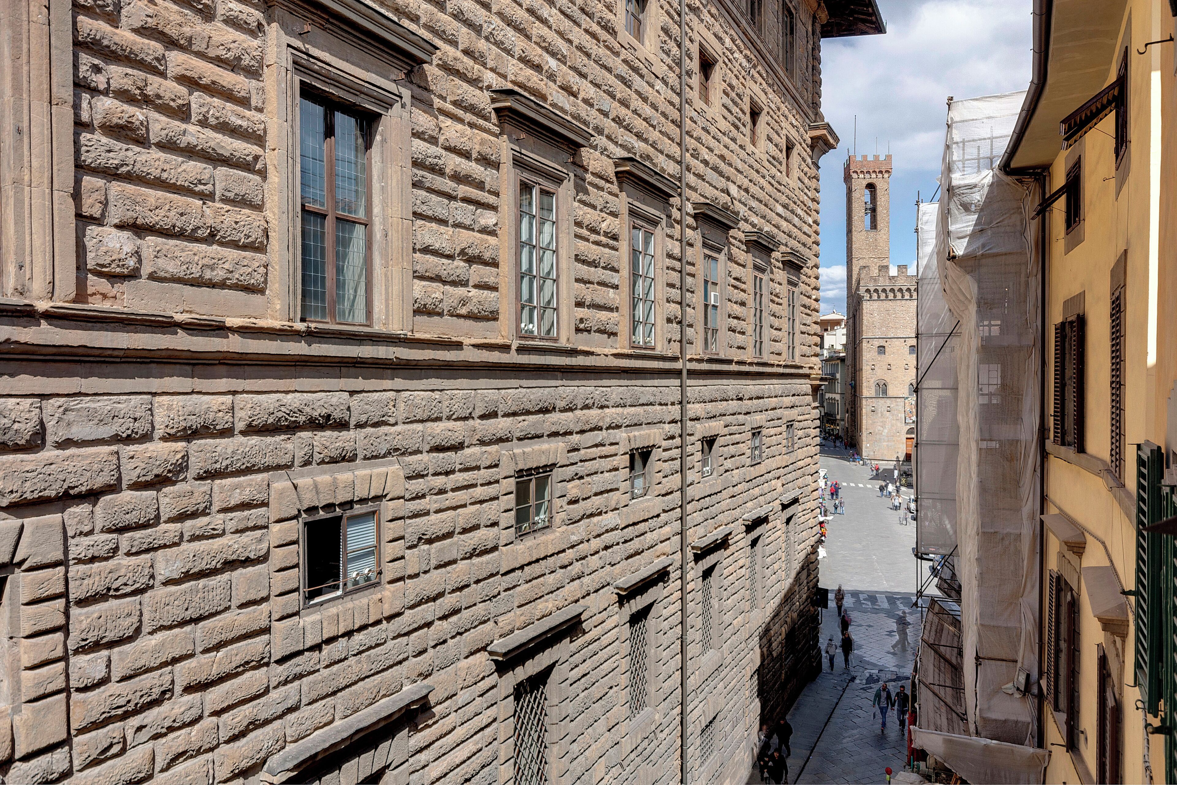 A narrow street with historic stone buildings, a tower in the distance, and a clear sky.