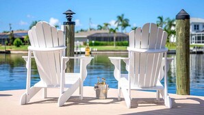 Two white Adirondack chairs facing a body of water with a small table and a bucket of bottles between them.