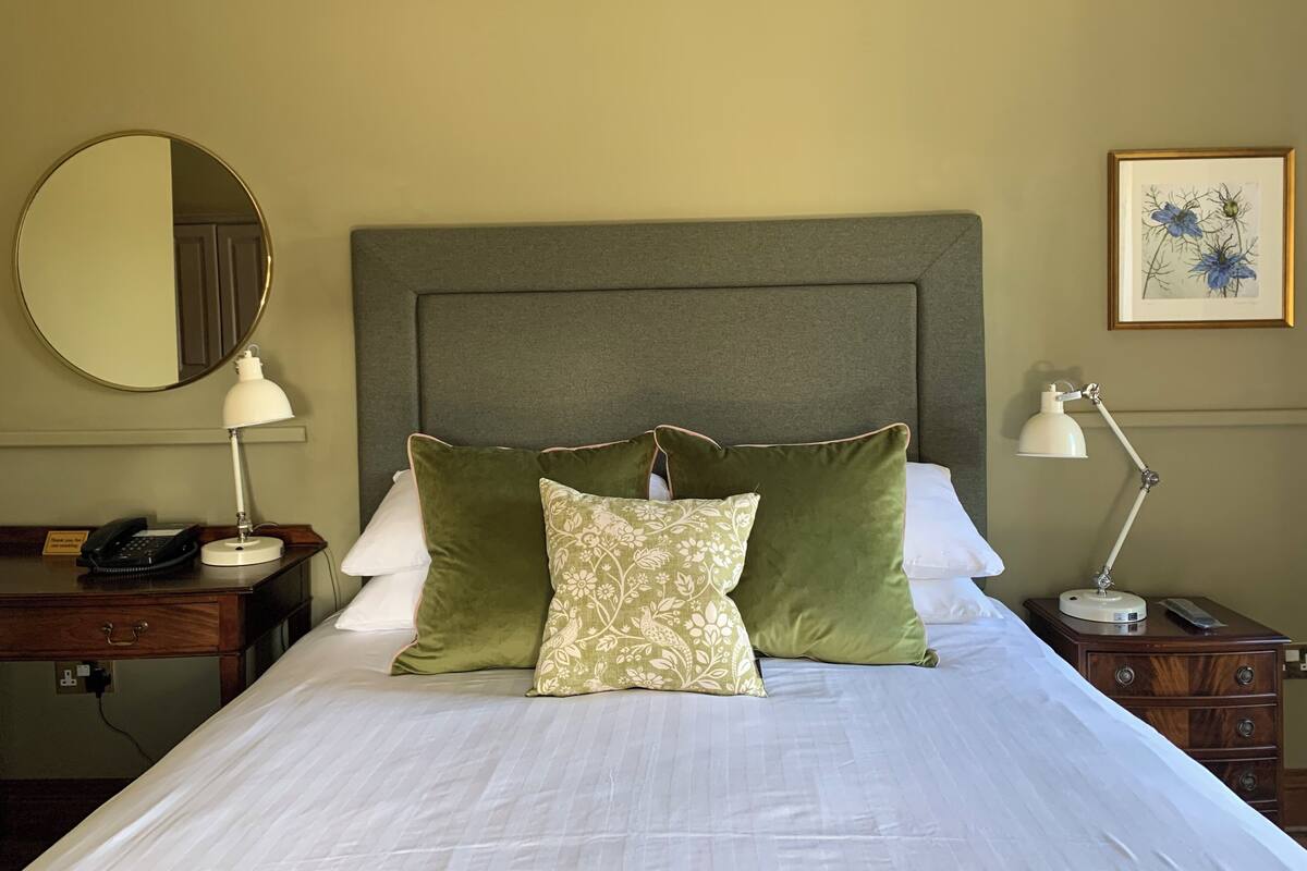 A neatly made bed with green and patterned pillows, a round mirror, a bedside lamp, and a framed botanical print on the wall.