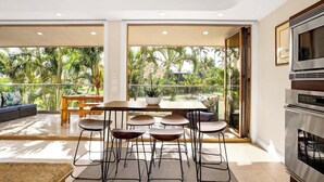 A modern kitchen with a large island, a dining area with a table and chairs, and a view of palm trees through large windows. - Aloha Villas # 4, Luxury Oceanfront Townhouse, Ste (Kihei)