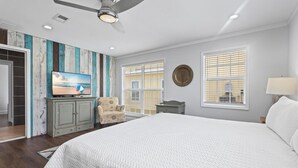 A bedroom with a large bed, a ceiling fan, a TV on a cabinet, a chair, and two windows with blinds.