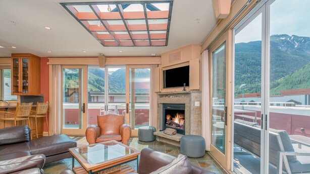 Un vaste espace de vie doté de canapés en cuir, d’une table basse en verre, d’une cheminée et d’une grande fenêtre offrant une vue sur les montagnes.