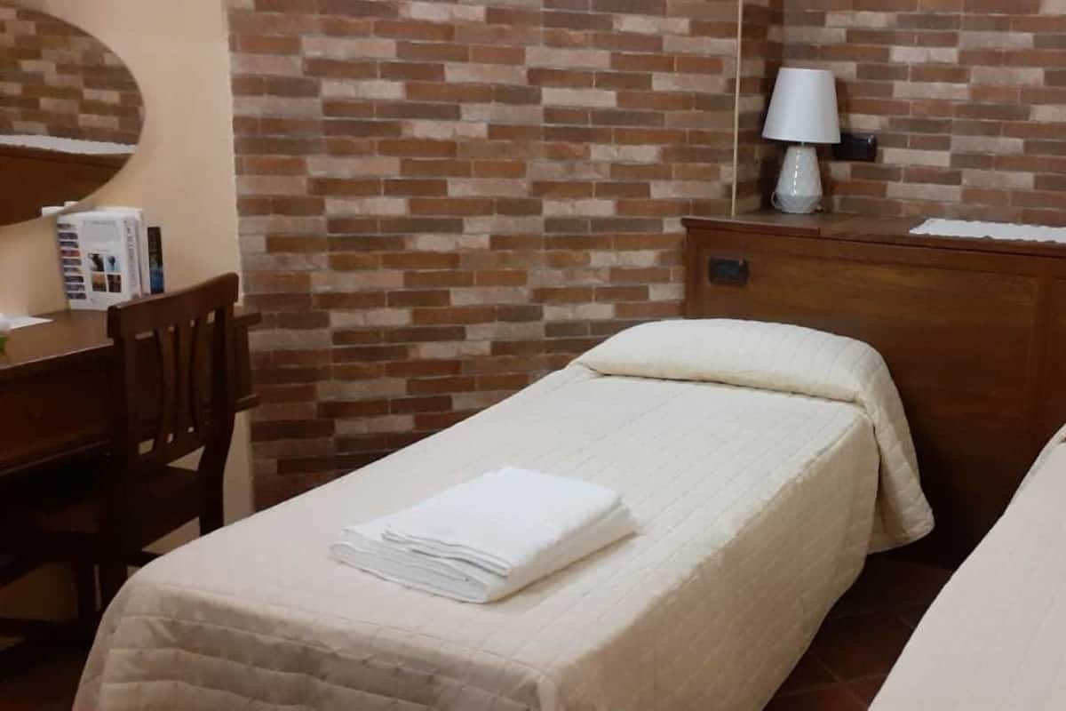 A hotel room with two beds, a wooden dresser, a desk with a chair, a mirror, and two wall-mounted lamps.