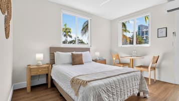 A hotel room with a bed, two bedside tables, a chair, a small table, and two windows with a view of palm trees.