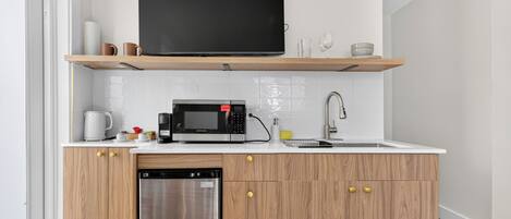 A compact kitchen with a built-in microwave, a stainless steel dishwasher, and a flat-screen TV mounted on a wooden shelf above the countertop.