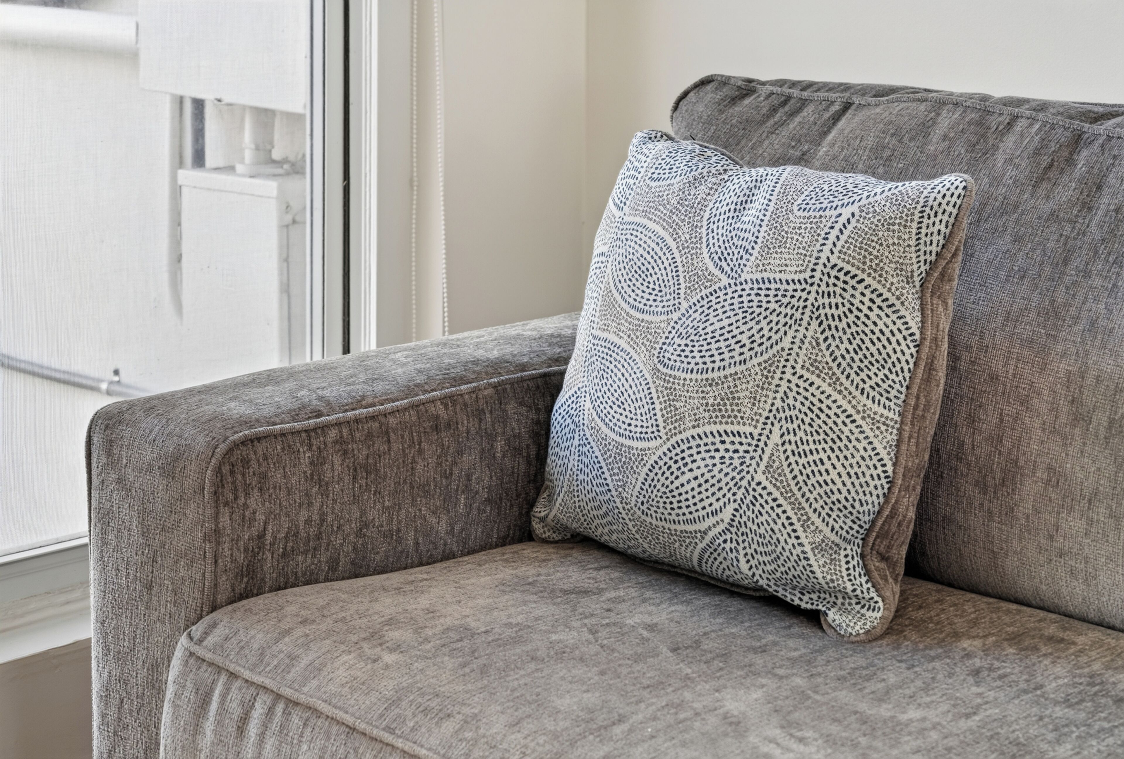 Un sofá tapizado en gris con tela texturizada y una almohada de punto blanco con un diseño circular.