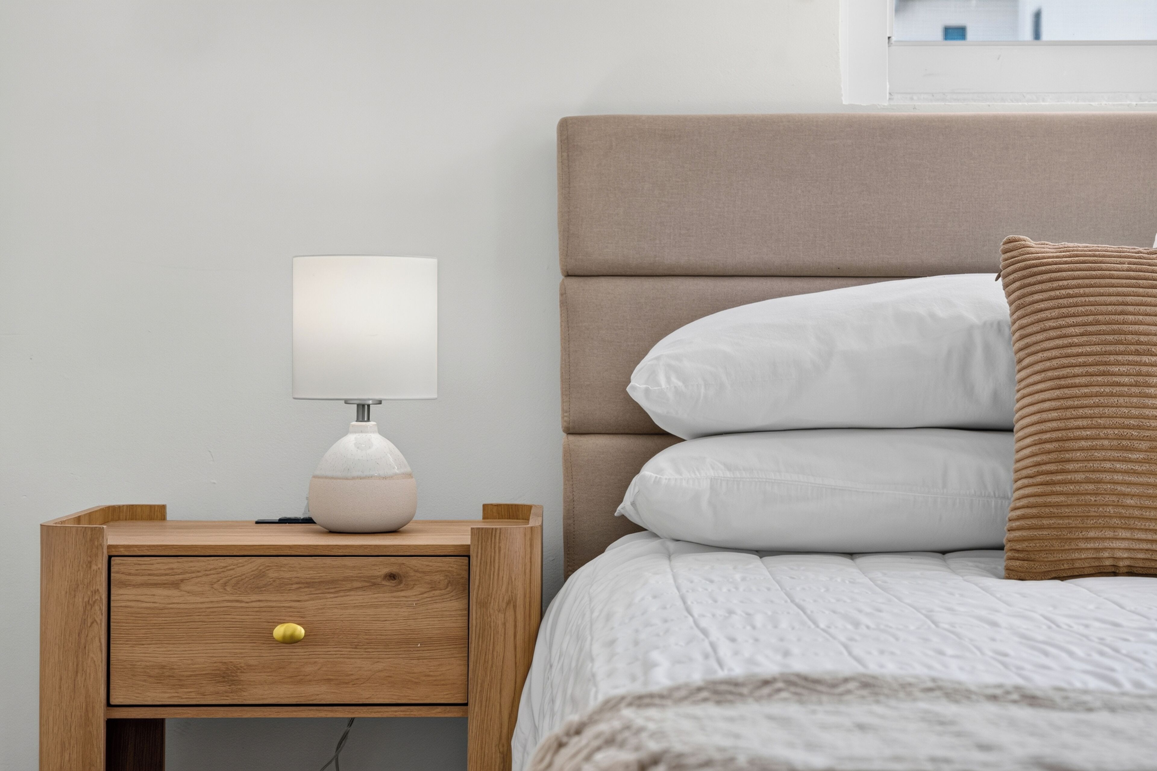 Ein ordentlich bezogenes Bett mit weißer Bettwäsche und einem braunen Kopfteil, ein Nachttisch mit einer Lampe und eine Kommode mit einem gelben Knauf.