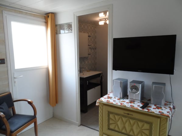 A room with a television mounted on the wall, a small table with speakers, a chair, and a door leading to a bathroom.