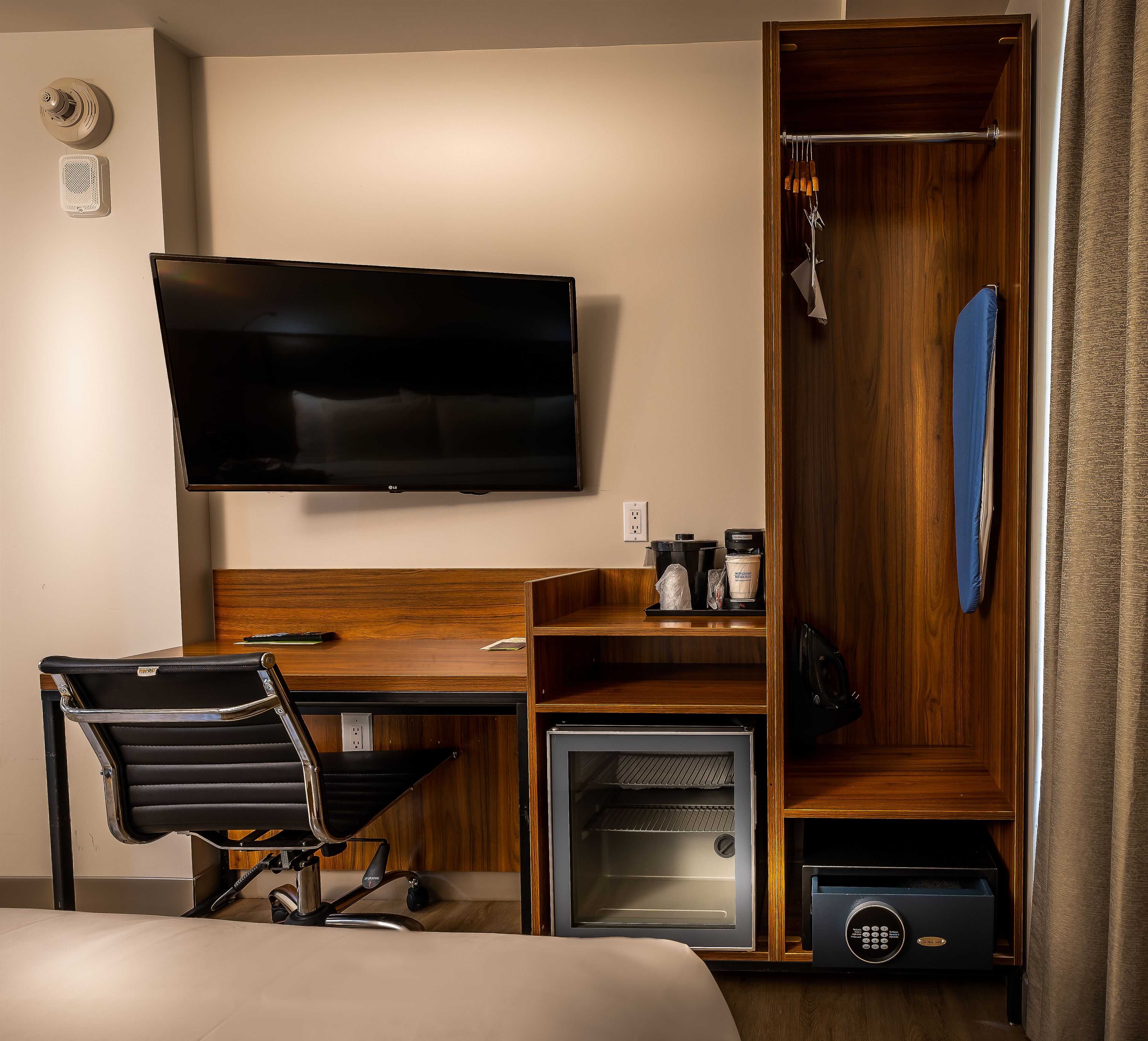 A hotel room with a flat-screen TV, a desk with a chair, a minibar, a coat rack, and a wardrobe.