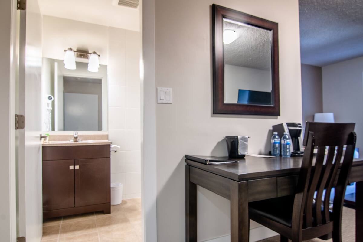 Une salle de bain avec un lavabo, un miroir et un meuble-lavabo. On y trouve une chaise en bois et une table avec une lampe.