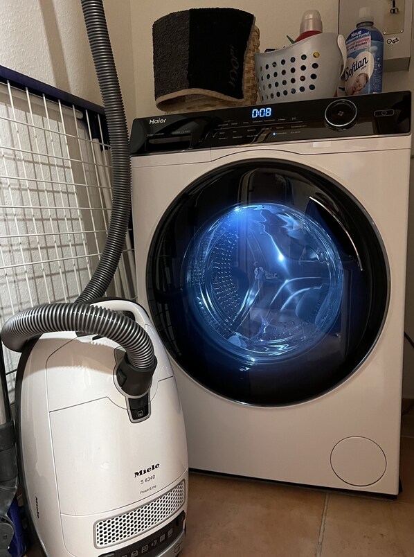 A front-loading washing machine with a digital display, a vacuum cleaner, and a laundry basket. - Exquisite apartment on one of the most beautiful beaches in Germany (Prerow)