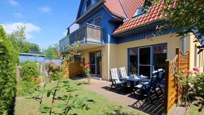 Een huis met twee verdiepingen, een balkon, een terras met stoelen en een tafel, en een tuin met bloemen.