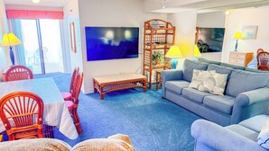 A living room with a blue sofa, a wooden coffee table, wicker chairs, and a TV mounted on the wall.