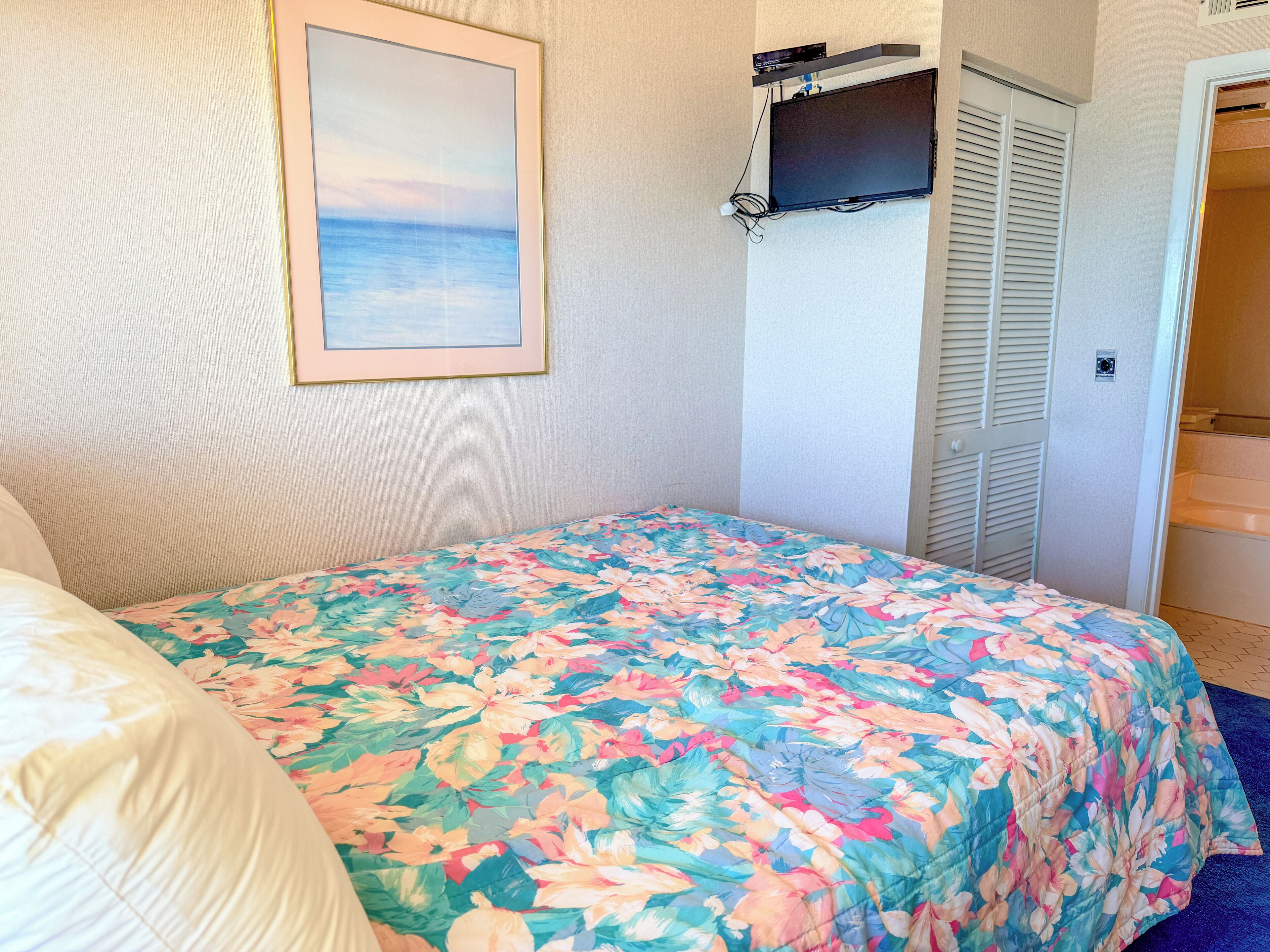 A bedroom with a floral bedspread, a framed picture on the wall, and a television mounted above the bed.
