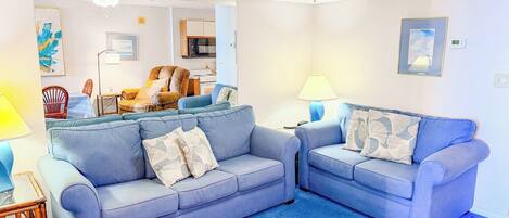 A living room with two sofas, a coffee table, and a chair. There is a ceiling fan and a lamp on a side table.