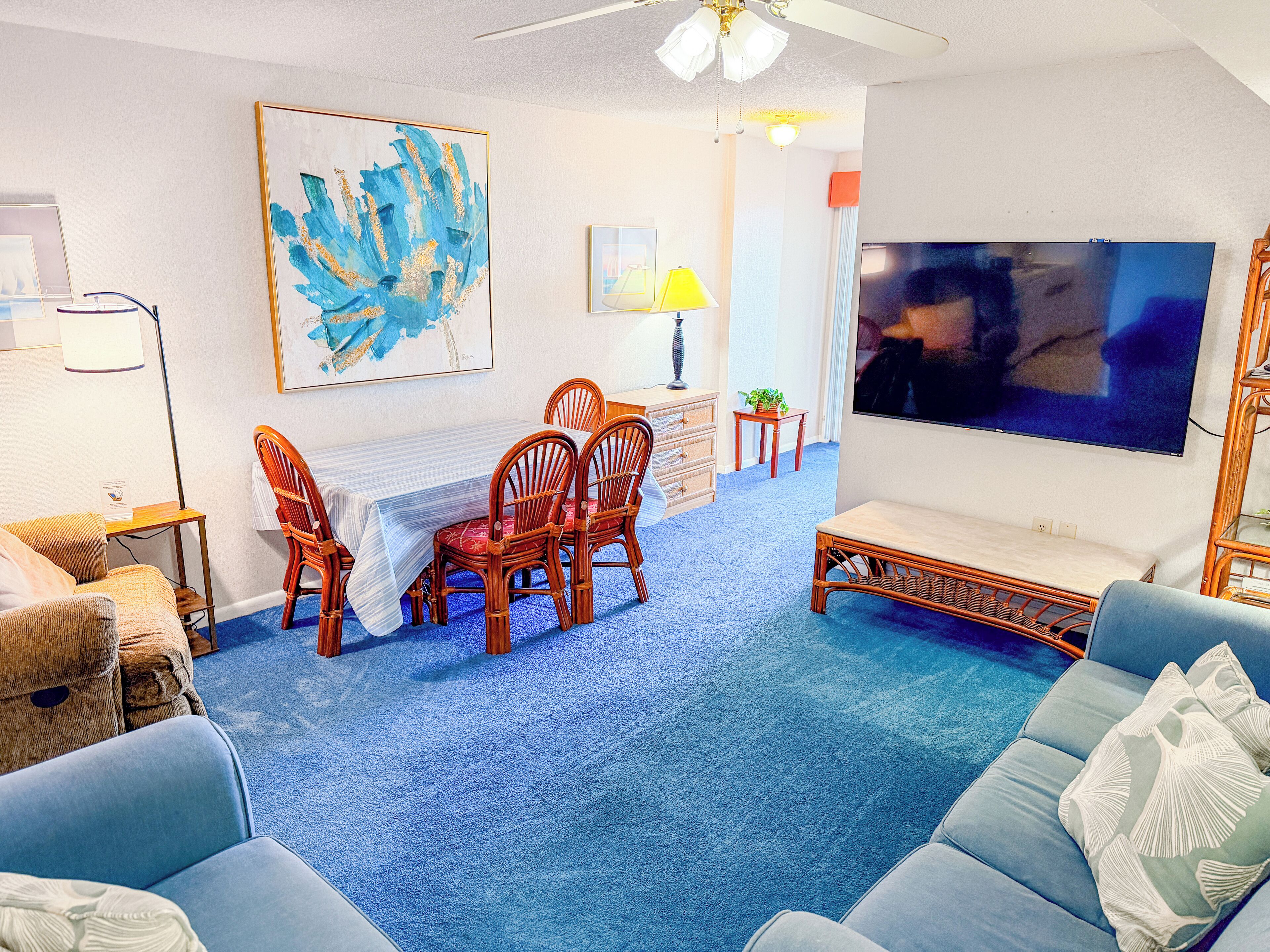 Una sala de estar con alfombra azul, una mesa de comedor con sillas, un sofá, un televisor y obras de arte en las paredes.