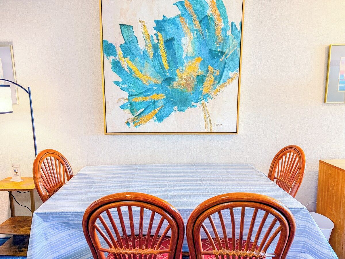 A dining area with a table set for four, wooden chairs, and a painting on the wall.