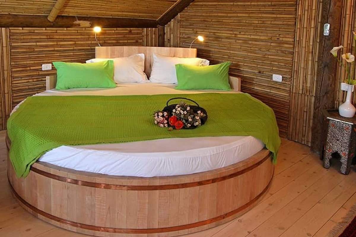 Un lit rond en bois, dans une pièce aux murs recouverts de bambou et orné d’une couverture verte.