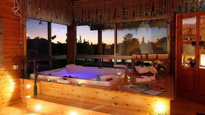 Een houten chalet met een groot houten bad, een raam met uitzicht op de zonsondergang, en een gezellig interieur met een kleine tafel en stoelen.
