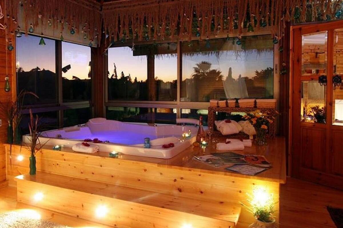 Une cabane en bois dotée d’une grande baignoire en bois, d’une fenêtre offrant une vue sur le coucher de soleil et d’un intérieur chaleureux avec une petite table et des chaises.