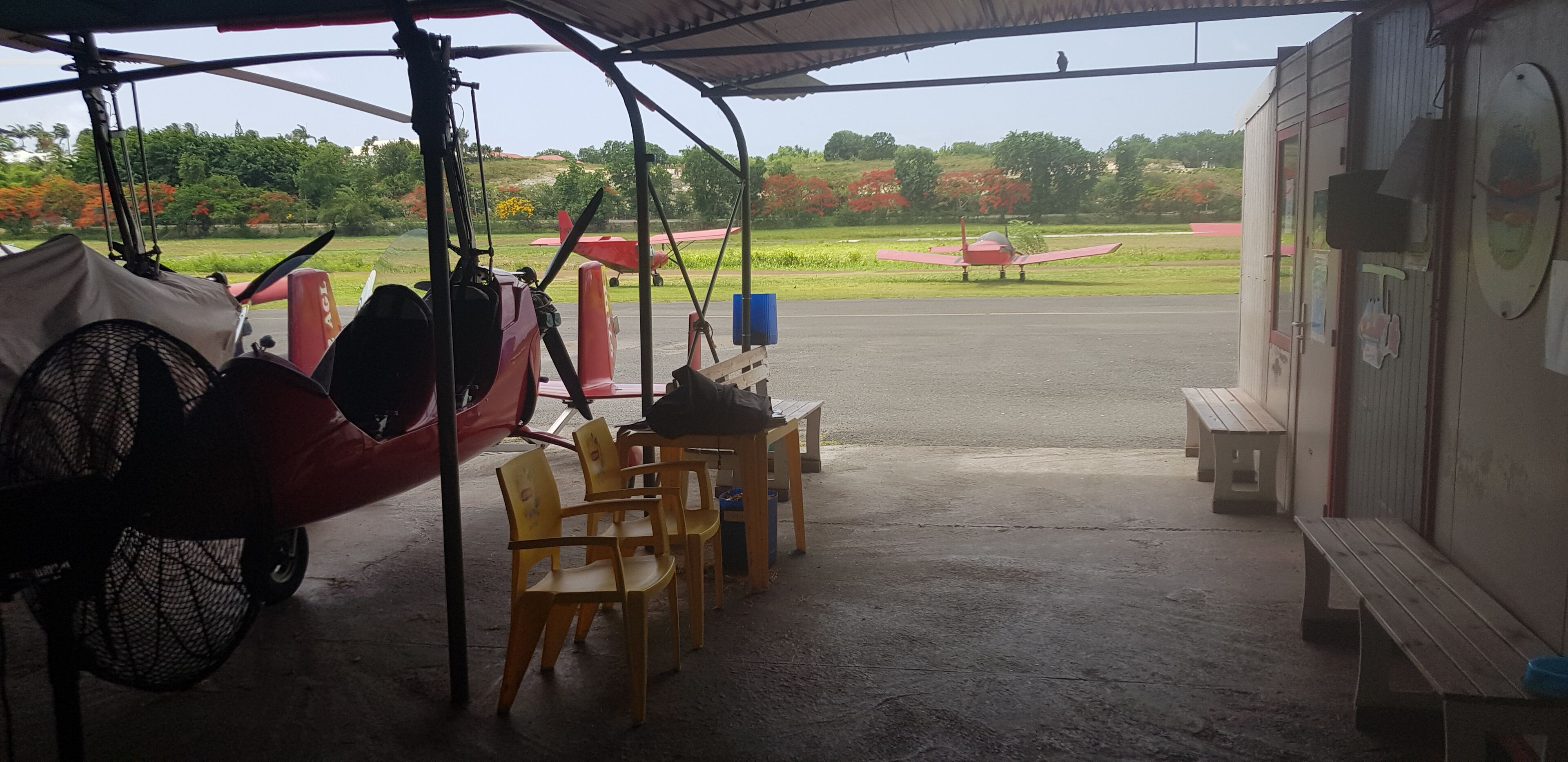 A covered area with chairs and a table, a red airplane on the ground, and a building in the background.