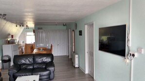 Ett vardagsrum med en svart lädersoffa, ett matbord med stolar, en köksdel med ett kylskåp och en stor TV som är monterad på väggen.