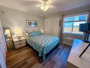 A bedroom with a bed, a nightstand, a lamp, a ceiling fan, a window with blinds, and a view of the outdoors.