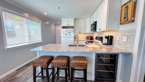 A modern kitchen with a marble countertop, white cabinets, and a built-in oven. There are three wicker bar stools and a window with blinds. - Inlet Point Villas #2A! Relax & Recharge in Cherry Grove’s Most Peaceful Corner (North Myrtle Beach)