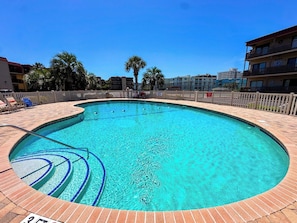 A clear blue swimming pool with steps leading into it, surrounded by a brick deck and a wooden fence.
