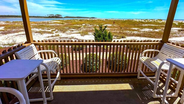 A covered outdoor seating area with two white plastic chairs and a table, overlooking a beach with sand dunes and a clear blue sky. - Inlet Point Villas #2A! Relax & Recharge in Cherry Grove’s Most Peaceful Corner (North Myrtle Beach)